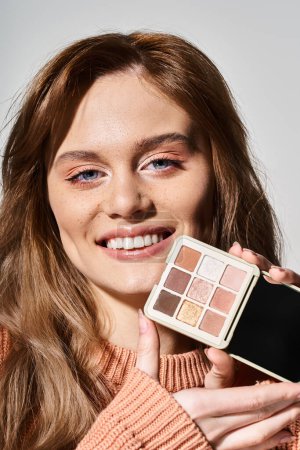 Téléchargez les photos : Plan beauté d'une femme souriante tenant une palette de maquillage pêche près du menton sur fond de studio gris - en image libre de droit