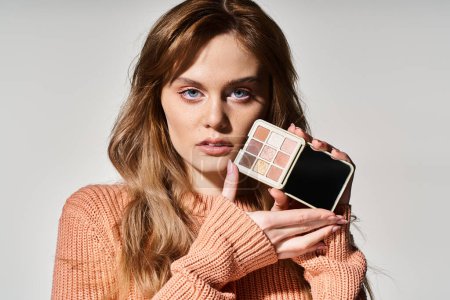 Beauty portrait of woman holding peach makeup palette near chin looking at camera on grey background
