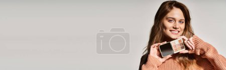 Foto de Foto de belleza de mujer sonriente con paleta de maquillaje de melocotón cerca de la barbilla sobre fondo gris estudio, pancarta - Imagen libre de derechos
