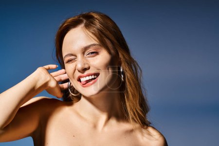 Smiling woman making funny winky face with tongue, touching her ear on dark blue background
