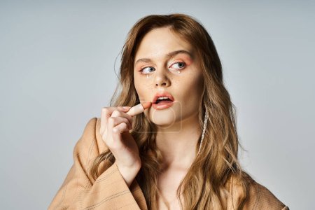 Attractive woman with face jewels in beige jacket, applying lipstick and looking away, peach fuzz