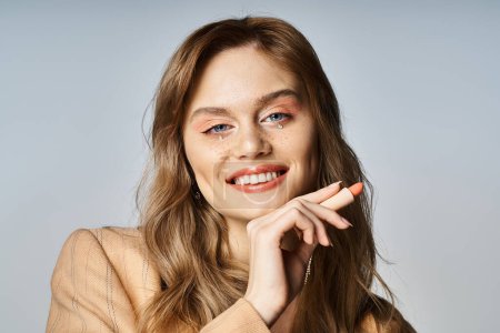 Téléchargez les photos : Femme souriante avec des bijoux visage en veste beige, tenant du rouge à lèvres de couleur pêche sur fond gris - en image libre de droit