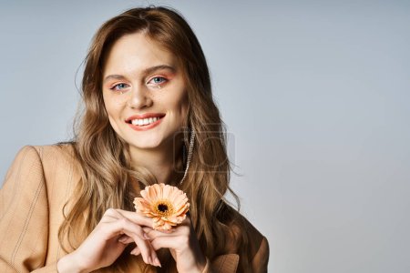 Foto de Mujer atractiva sonriente mirando a la cámara y sosteniendo la margarita, usando maquillaje de melocotón, joyas de la cara - Imagen libre de derechos