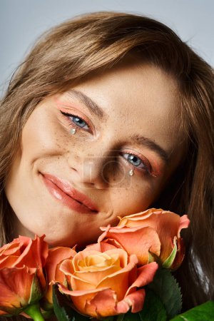 Closeup photo of smiling woman with peach makeup, face jewels and freckles, holding roses near cheek