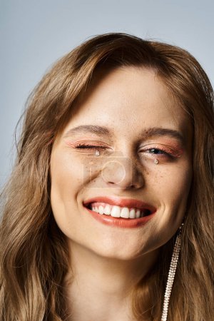 Foto de Primer plano de mujer sonriente guiñando con maquillaje de melocotón, joyas de la cara y pecas sobre fondo gris - Imagen libre de derechos