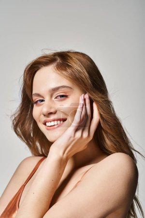 Téléchargez les photos : Jeune jolie femme avec une peau parfaite souriant à la caméra toucher le visage, sur fond de studio gris - en image libre de droit