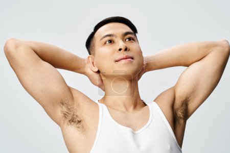 A handsome Asian man in a white tank top holds his hands behind his head in a serene and powerful pose in a grey studio.