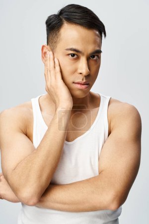 Photo for A handsome Asian man in a white tank top raises his hand to his face in a contemplative gesture inside a grey studio. - Royalty Free Image