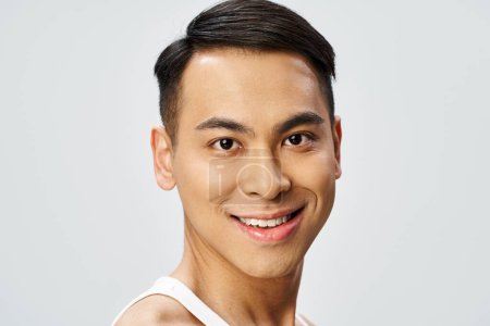 Ein gutaussehender asiatischer Mann in weißem Tank-Top lächelt in einem grauen Studio freundlich in die Kamera.