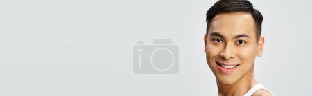 Téléchargez les photos : Un bel homme asiatique dans un débardeur blanc sourit sereinement à la caméra dans un décor de studio gris. - en image libre de droit