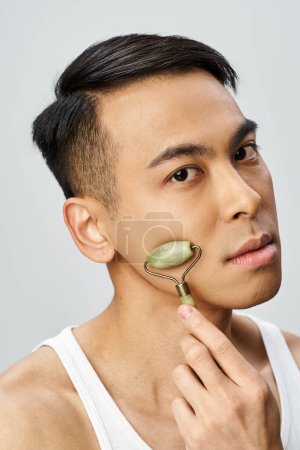 Guapo asiático hombre usando jade roller en su piel en un gris estudio.