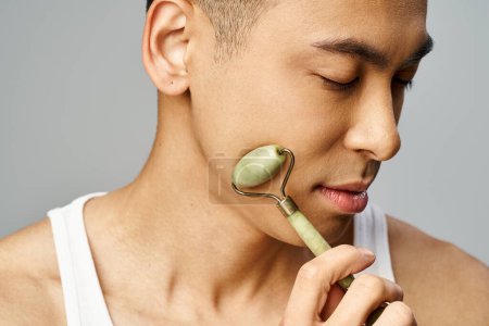 A handsome Asian man in a grey studio holding a jade roller