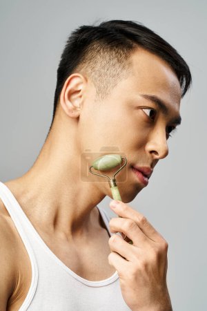 Handsome Asian man carefully using jade roller in a modern grey studio setting.