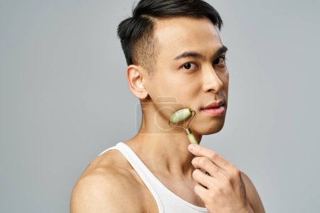 Asian man using jade roller, focusing on grooming and self-care routine in a grey studio setting.