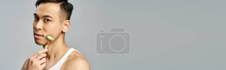 Photo for Handsome Asian man diligently using jade roller in a serene grey studio, a skincare routine evident in his meticulous care. - Royalty Free Image