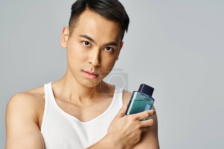 Foto de Un hombre asiático guapo en una camiseta delicadamente sostiene una botella de perfume en un estudio gris. - Imagen libre de derechos