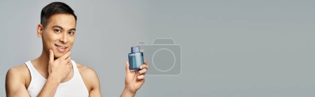 Photo for A handsome Asian man holds a bottle of perfume in a grey studio setting, banner - Royalty Free Image