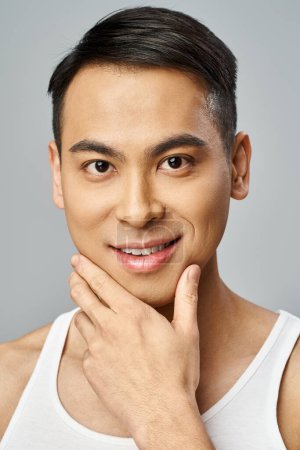 Schöner asiatischer Mann, in einem Tank-Top, posiert selbstbewusst in einem grauen Studio während einer Hautpflege-Routine.