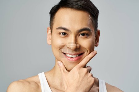 Ein gutaussehender asiatischer Mann mit einem Lächeln im Gesicht, der in einem grauen Studio Glück und Zuversicht ausstrahlt, während er Pflegeprodukte verwendet.