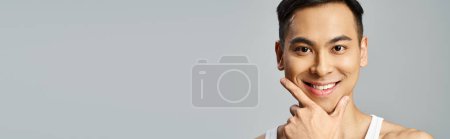 Foto de Un hombre asiático guapo con una camiseta blanca posa con elegancia, mostrando su rutina de cuidado de la piel en un sereno estudio gris. - Imagen libre de derechos