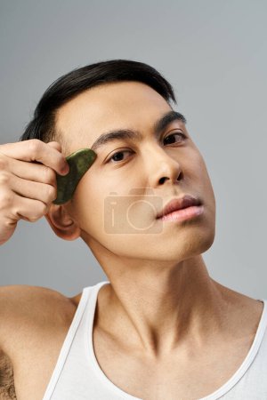 Schöner asiatischer Mann in einem grauen Studio hält eine grüne Gua-sha in einem grauen Studio