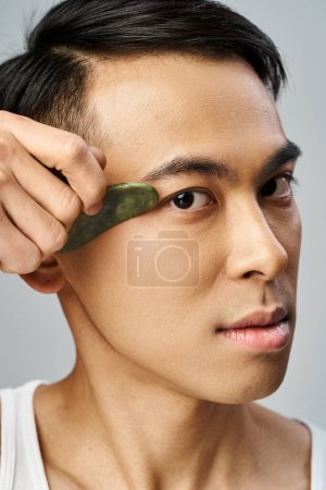 A handsome Asian man in a grey studio holds a green gua sha in his hand on grey