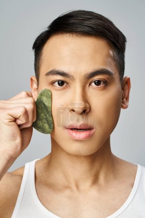 Photo for Asian man using green gua sha on his face in a grey studio setting. - Royalty Free Image