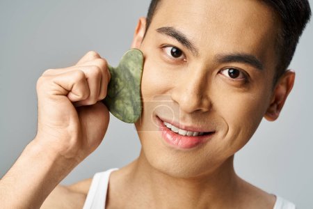 Foto de Un hombre asiático guapo sostiene una piedra verde en su cara en un estudio gris, mostrando su rutina de cuidado de la piel. - Imagen libre de derechos