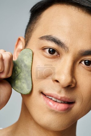 Photo for Handsome Asian man in grey studio holding gua sha to face for skincare routine. - Royalty Free Image
