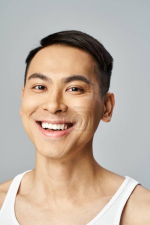 A handsome Asian man is beaming with a smile, exuding warmth and happiness in a grey studio while using skin care products.