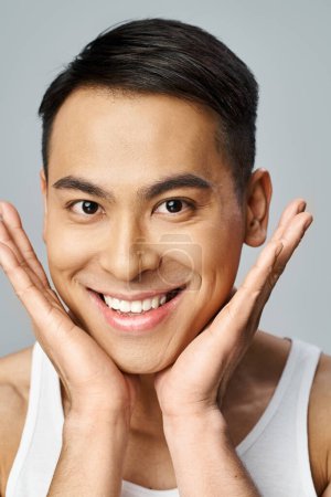 A handsome Asian man is smiling brightly in a grey studio after using skincare products.
