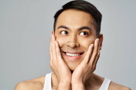 Foto de Hombre asiático guapo con una sonrisa serena, tocándose suavemente la cara en una rutina calmante para el cuidado de la piel en un estudio gris. - Imagen libre de derechos