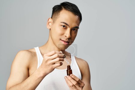 Téléchargez les photos : Beau asiatique dans un débardeur tenant une bouteille avec du sérum dans un décor de studio gris. - en image libre de droit