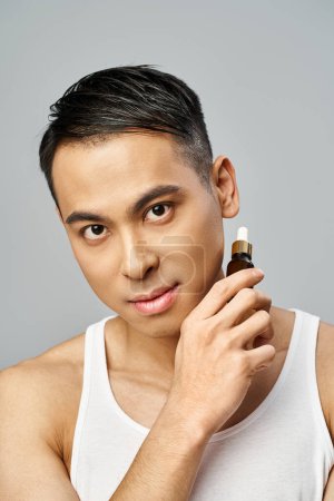 A handsome Asian man in a white tank top calmly holds a bottle with serum in a grey studio setting, focused on hydrating.