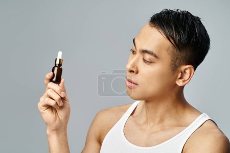 Téléchargez les photos : Un bel homme asiatique dans un studio gris tient délicatement une bouteille d'huiles essentielles, incarnant détente et autosoin. - en image libre de droit