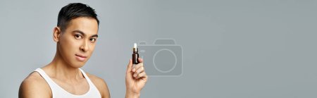 Téléchargez les photos : Un bel homme asiatique dans un studio gris tient délicatement une bouteille d'huiles essentielles, incarnant détente et autosoin. - en image libre de droit