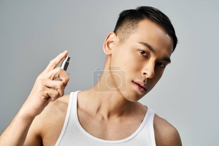Téléchargez les photos : Un bel homme asiatique dans un débardeur pulvérisant son parfum luxueux dans un studio gris. - en image libre de droit
