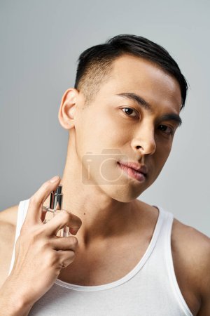 Schöner asiatischer Mann in weißem Tank-Top, der Parfüm in einem grauen Studio versprüht.