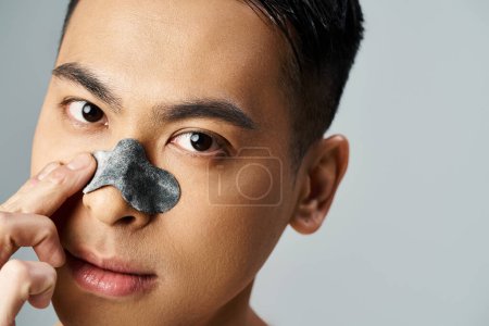 A handsome Asian man wearing a nose patch in a beauty and skin care routine, set in a grey studio.