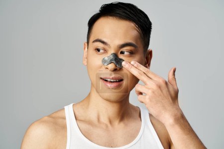 Téléchargez les photos : Un homme asiatique avec un masque facial en tenant sa main sur son visage dans un portrait de routine beauté et soins de la peau dans un studio gris. - en image libre de droit
