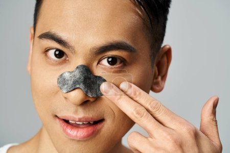 Ein gutaussehender asiatischer Mann in einem grauen Studio mit Schönheitsnasenpflaster, Hautpflege-Routine.