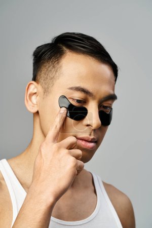 Handsome Asian man in a white tank top holds a pair of black under eye patches in a grey studio setting during a beauty routine.