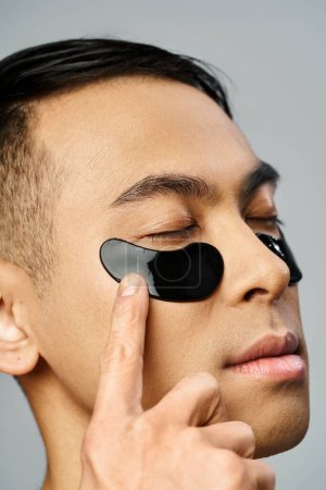 Asian man with a striking black eye patch enhancing his skin during a beauty routine in a grey studio.