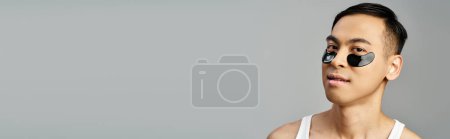 Porträt eines gut aussehenden asiatischen Mannes, der während seiner Hautpflege-Routine in einem grauen Studio eine Augenklappe wippt.