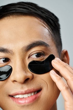 Schöner asiatischer Mann trägt in einem grauen Studio schwarze Augenklappen für seine Hautpflege auf.