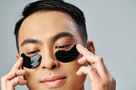 Un bel homme asiatique tenant deux cercles noirs jusqu'aux yeux dans un studio gris, se concentrant sur sa routine beauté et soins de la peau.
