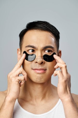 Handsome Asian man holding two black circles over his eyes in a beauty and skincare routine in a grey studio.