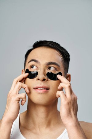 Hombre asiático guapo con parches debajo de los ojos que participan en una rutina de belleza y cuidado de la piel en un estudio gris.