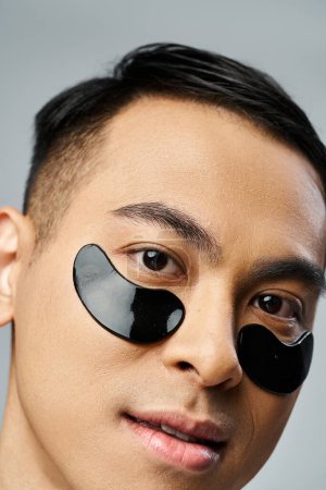 Hombre asiático guapo con parches debajo de los ojos durante la rutina de belleza y cuidado de la piel en estudio gris.