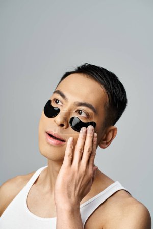 Handsome Asian man with black eye patches, in a beauty and skin care routine, in a grey studio setting.